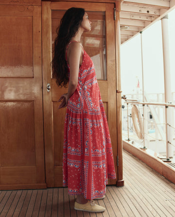 A woman stands in profile on a wooden deck near a door, wearing THE GREAT. SU26's The Drop Waist Dress in Red Sailboat Bandana, with hands behind her back and looking ahead.