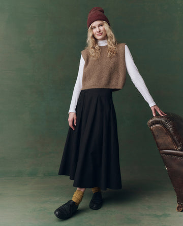 A person wears THE GREAT. F25 D2 The Brushed Melange Sweater by Doe over a white shirt, teamed with a black skirt, yellow socks, black shoes, and a red beanie, standing beside a brown armchair against a green backdrop.