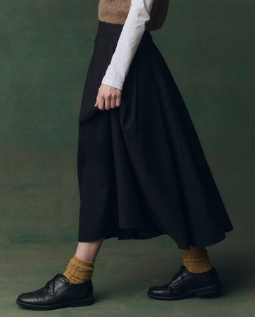 A person in a white long-sleeve top, THE GREAT. F25 D2 The Regency Skirt in black, mustard yellow socks, and black shoes walks against a green background.