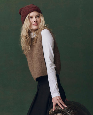 A woman with long blonde hair wears THE GREAT.'s The Brushed Melange Sweater by Doe over a white long-sleeve shirt, paired with a maroon knit beanie and black skirt, leaning on a brown leather chair.