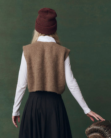 Wearing THE GREAT. F25 D2’s Doe Brushed Melange Sweater, a white long-sleeve shirt, black skirt, and burgundy beanie, a person stands facing away against a dark green background.