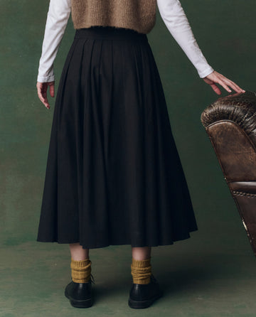 A person stands with their back to the camera, wearing THE GREAT. F25 D2's The Regency Skirt in black, yellow socks, black shoes, a structured white cotton long-sleeve top, and a brown vest beside a brown armchair.