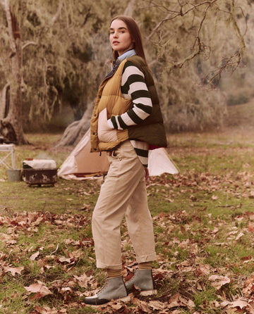 In a forested area, an adventurer embodies the spirit of exploration wearing technical attire: a striped sweater, The Down Polar Vest in Appalachian Colorblock from THE GREAT. FALL 24 TGO, khaki pants, and boots. Tents are scattered throughout the landscape while moss-covered trees create a tranquil atmosphere. Ideal for eco-friendly explorers, this vest features responsibly sourced down for superior warmth and sustainability.