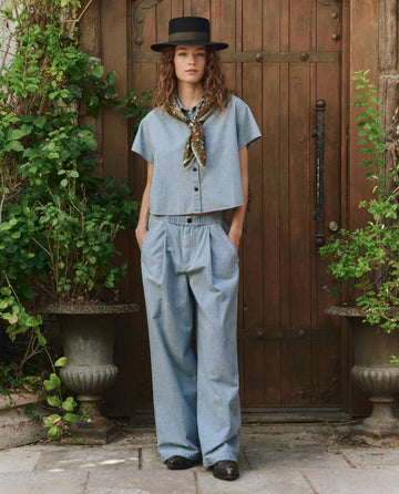 A person stands before a wooden door wearing stylish attire, featuring THE GREAT. PF25's The Valor Top in Boiler Wash, a wide-brimmed hat, wide-leg trousers, a patterned scarf, and black shoes.