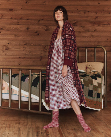 Dressed in The Ruffle Tank Night Dress in Bordeaux Plaid by THE GREAT. HOL 24 SLEEP, a person stands gracefully in a wooden room with a metal bed frame and patchwork quilt, exuding the cozy elegance reminiscent of a classic night dress.