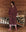 A person in The Plush Fleece Robe by THE GREAT. HOL 24 SLEEP in Fireside Plaid and cozy socks stands on a wooden floor beside a metal bed with a quilt.