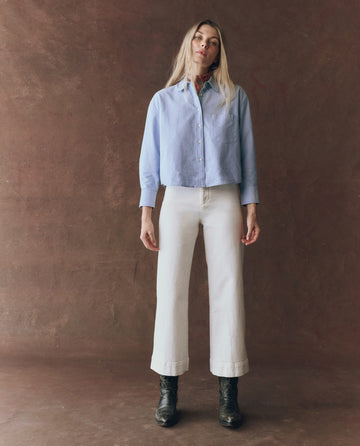 A woman faces forward against a brown background, wearing THE GREAT. F25 D1 The Cropped Oxford Shirt in Classic Blue, white wide-leg pants, and black boots.