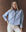 A woman stands against a brown background wearing THE GREAT. F25 D1's The Cropped Oxford Shirt in Classic Blue, styled with white pants and a red bandana around her neck.
