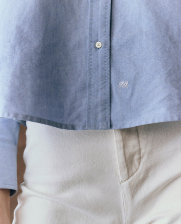 A person wears THE GREAT. F25 D1's Cropped Oxford Shirt in Classic Blue, styled with high-waisted off-white pants; photographed from chest to thigh to highlight the boxy, cropped fit.