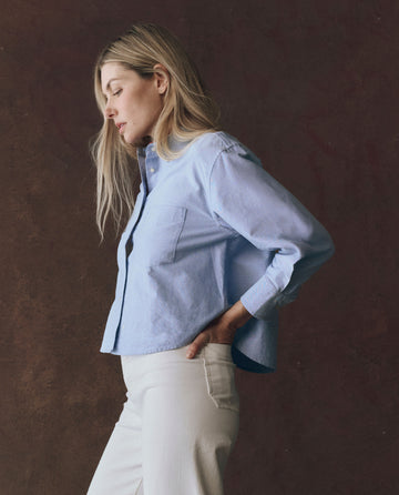 A woman with long blonde hair stands in profile against a brown background, wearing THE GREAT. F25 D1's The Cropped Oxford Shirt in Classic Blue and off-white pants.