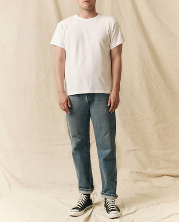 A person stands before a beige fabric backdrop, wearing THE GREAT. FALL 23 MEN The Men's Pure Knits Boxy Crew in Solid True White, paired with blue jeans and black sneakers.
