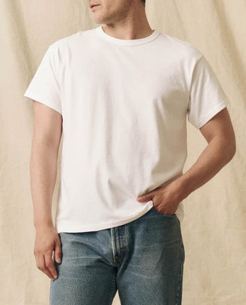 A person wears THE GREAT. FALL 23 MEN The Men's Pure Knits Boxy Crew, Solid -- True White, and blue jeans, standing against a beige fabric background.