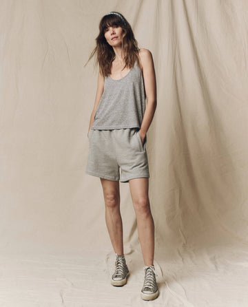 A woman stands with hands in pockets, wearing a gray tank top, THE GREAT. SU25 CORE KNITS The Victory Short. Solid -- Varsity Grey, and sneakers against a beige fabric backdrop.