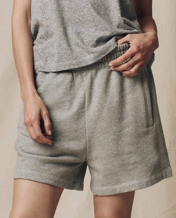 Someone wearing THE GREAT. SU25 CORE KNITS The Victory Short. Solid -- Varsity Grey with a matching tee stands against a neutral background, holding the waistband—perfectly highlighting this performance wear ensemble.