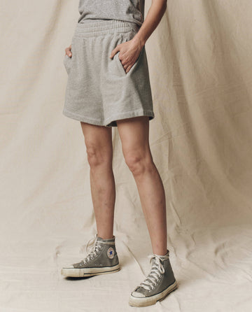 The lower half of a person is shown in front of a beige fabric backdrop, wearing THE GREAT. SU25 CORE KNITS The Victory Short. Solid -- Varsity Grey, a gray t-shirt, and gray high-top sneakers.