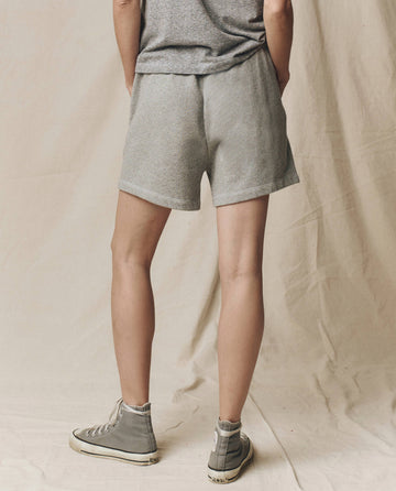 Person stands with their back to the camera, wearing a grey t-shirt and THE GREAT. SU25 CORE KNITS The Victory Short. Solid -- Varsity Grey, paired with grey high-top sneakers against a neutral fabric backdrop.