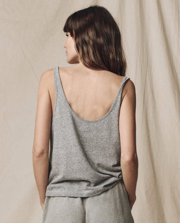 A woman with her hair down faces away, wearing THE GREAT. SU25 CORE KNITS The Scoop Neck Tank in heather grey, paired with matching pants. A neutral backdrop emphasizes the modern style of this women's clothing piece.