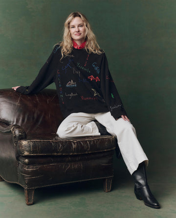 A woman with wavy blond hair sits on a worn leather chair wearing THE GREAT. F25 D2's Slouch Sweatshirt in Almost Black with Golden State Embroidery, white pants, and black boots, against a green background.