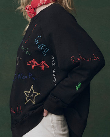 Someone wears THE GREAT.'s The Slouch Sweatshirt, Embroidered -- Almost Black with Golden State embroidery, light pants, and a red bandana around their neck, standing against a dark green background.