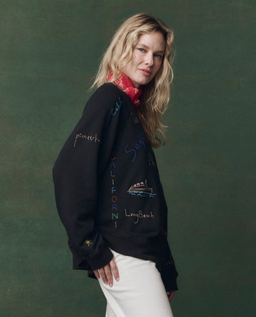 A woman with long blonde hair wears THE GREAT. F25 D2's The Slouch Sweatshirt in Almost Black with Golden State Embroidery, paired with a red scarf and white pants, standing against a green background.