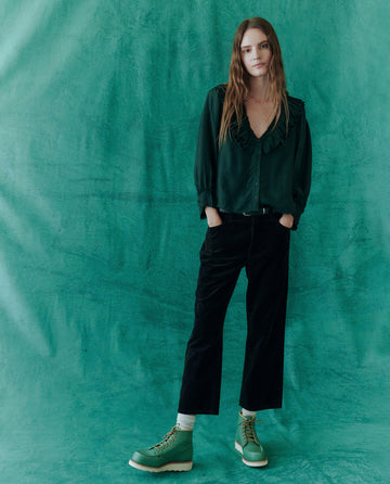 A person with long hair is wearing a dark ruffled blouse, The Velvet Wayne Jean by THE GREAT. HOL 24 in black, and green boots, standing against a green textured background.