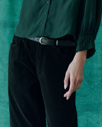 A person in a green blouse with buttons and The Velvet Wayne Jean in black, set against a lush green background, channels the elegance of THE GREAT. HOL 24.