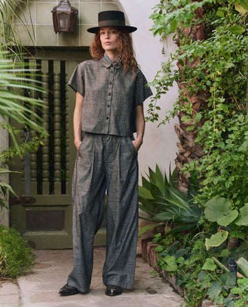 A person in THE GREAT. PF25 The Valor Top—Smoke Wash, a relaxed gray top and pants set—stands in a garden courtyard before a green door, embodying effortless style with comfort and elegance.