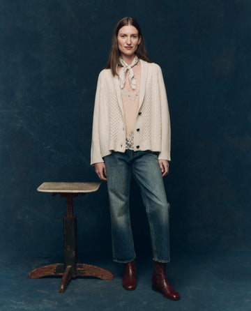 A woman stands indoors beside a small wooden table, wearing THE GREAT. F25 COLLECTORS The Cashmere Cable Lodge Cardigan in cream, paired with jeans, maroon boots, and a scarf, set against a dark blue backdrop.
