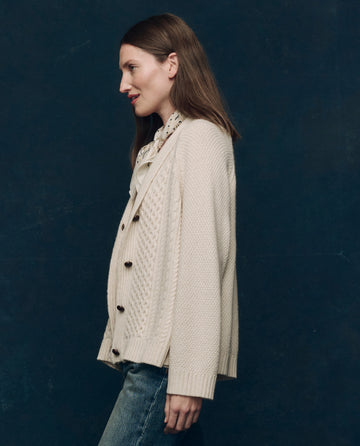 A woman with straight brown hair wears THE GREAT. F25 COLLECTORS' The Cashmere Cable Lodge Cardigan in cream and a light scarf, standing in profile against a dark background.