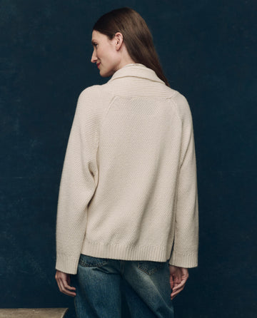 A woman faces away, wearing THE GREAT. F25 COLLECTORS' The Cashmere Cable Lodge Cardigan in cream with leather buttons and blue jeans against a dark background.