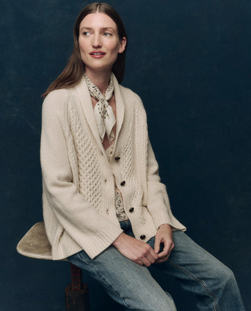 A woman with long brown hair sits on a stool, wearing THE GREAT. F25 COLLECTORS The Cashmere Cable Lodge Cardigan in cream, paired with a light patterned scarf and blue jeans, against a dark blue background.