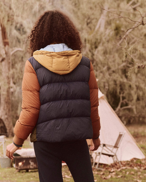 A person with curly hair, clad in the lively Appalachian Colorblock version of The Down Polar Puffer from THE GREAT. FALL 24 TGO, stands near a tent amidst the majestic expanse of The Great Outdoors.