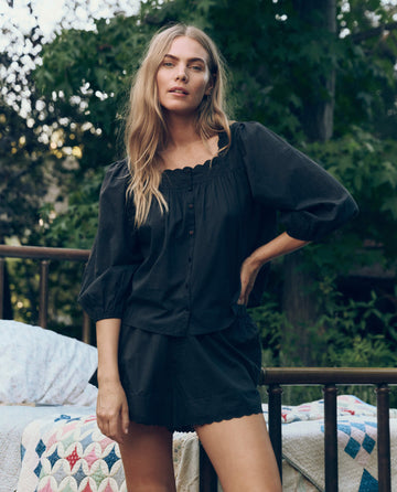 A woman with long blonde hair wears THE GREAT. CORE SLEEP’s The Eyelet Tap Short in black, standing outdoors near a bed with a patterned quilt, surrounded by trees and greenery.