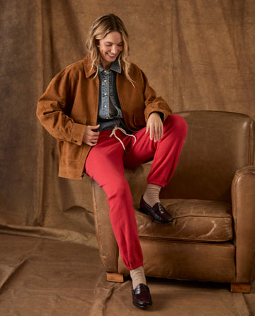 A woman wears a brown jacket over a denim shirt and red The Fleece Stadium Sweatpant while sitting on a leather chair.