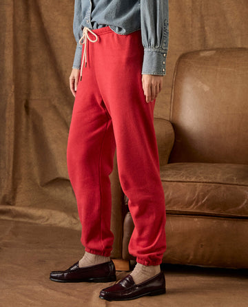 A person wearing red THE GREAT. H26 FLEECE BACK KNITS, The Fleece Stadium Sweatpant in Campfire with a drawstring waist and dark leather loafers.