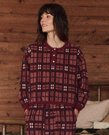 A person in The Plush Fleece Henley Sleep Sweatshirt, part of THE GREAT. HOL 24 SLEEP collection in Fireside Plaid, stands indoors against a wooden wall, gazing to the side.