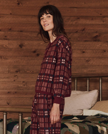 A person wearing The Plush Fleece Henley Sleep Sweatshirt in Fireside Plaid by THE GREAT. HOL 24 SLEEP stands in a room with wood-paneled walls and a bed.