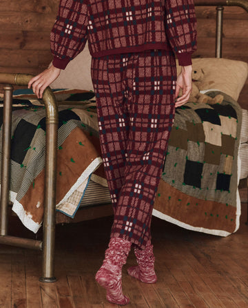 Wearing The Plush Fleece Pajama Pant in Fireside Plaid by THE GREAT. HOL 24 SLEEP, a person walks by a metal-framed bed covered with cozy patchwork quilts in a rustic wooden room.