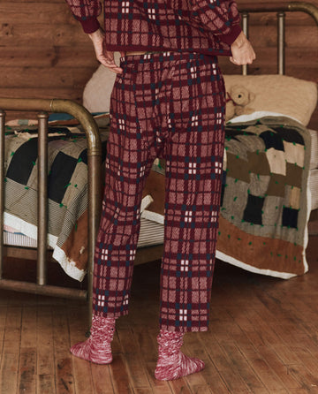 Dressed in The Plush Fleece Pajama Pant in Fireside Plaid from THE GREAT. HOL 24 SLEEP, a person stands by a bed adorned with a patchwork quilt, enveloped in the cozy warmth of a wooden room.