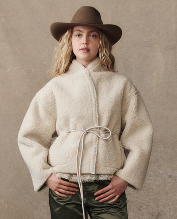 A person in THE GREAT. H25 COLLECTION's Reversible Drover Coat -- Malt, featuring cream fleece and a cord tie, with a tan wide-brim hat and dark green pants, stands against a plain beige background.