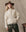 A person wearing THE GREAT. H25 COLLECTION’s The Reversible Drover Coat in Malt, a brown wide-brimmed hat, and dark green satin pants stands in front of a beige backdrop.
