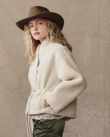 A person with long, wavy blonde hair wears a brown wide-brim hat, olive pants, and THE GREAT. H25 COLLECTION’s The Reversible Drover Coat in Malt, standing against a neutral background.