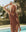 A woman wearing THE GREAT. SP25 SWIM's The Reversible Lace Up One Piece in Rust Tile Print and Hot Red stands by a pool outdoors, with trees and greenery in the background.