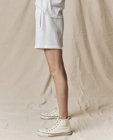 A person stands on a beige fabric background, wearing THE GREAT. SU25 CORE KNITS The Victory Short in Solid -- True White, paired with a white top and off-white high-top sneakers for all-day comfort.