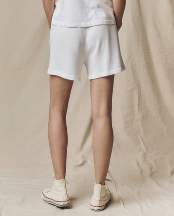 A person stands on a fabric background, shown from the back in THE GREAT. SU25 CORE KNITS The Victory Short Solid in True White, paired with a white top and high-top sneakers for a sporty yet effortlessly stylish look.