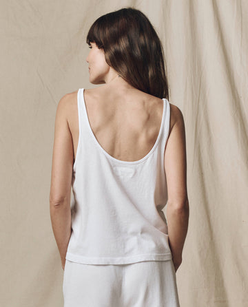 A woman faces away from the camera in front of a beige fabric backdrop, wearing THE GREAT. SU25 CORE KNITS The Scoop Neck Tank in True White and white bottoms.