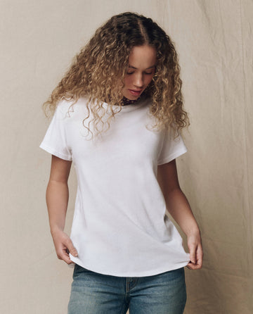 A person with curly hair wears THE GREAT. CORE KNITS The Slim Tee. Solid -- True White and blue jeans, standing against a neutral fabric backdrop, looking down and holding the shirt’s hem.