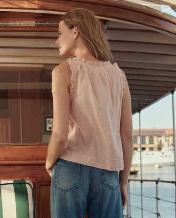 A person with long hair stands on a boat facing away, wearing THE GREAT. SU26 The Vignette Top in Petal and blue jeans, with water and buildings in the background.