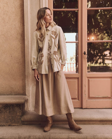 A woman in a complete beige outfit featuring The Floral Applique Suede Jacket in Tonal Desert Tan by THE GREAT. SP25 D1 stands near a wall and glass-paned door, wearing a long skirt and brown boots while gazing slightly to the side.