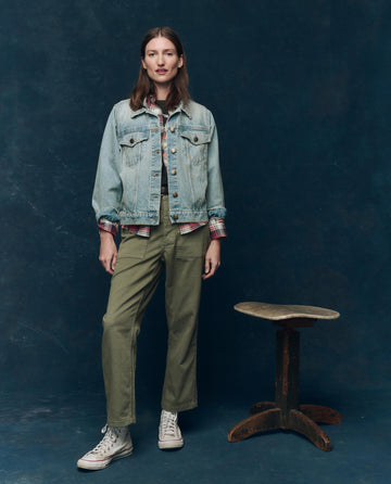 A person stands by a wooden stool, wearing THE GREAT. SP25 COLLECTORS The Slouchy Jean Jacket in Great Plains Wash over a plaid shirt, with olive green pants and white high-top sneakers, set against a dark blue backdrop.